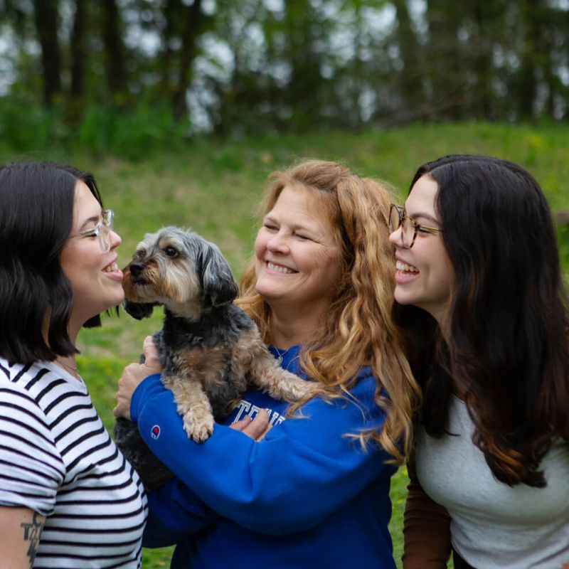 Candid photo of a woman with red hair smiling and holding a small dog outside in a yard. Her twin daughters stand on either side of her, smiling and laughing.