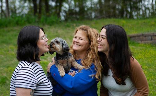 Candid photo of a woman with red hair smiling and holding a small dog outside in a yard. Her twin daughters stand on either side of her, smiling and laughing.