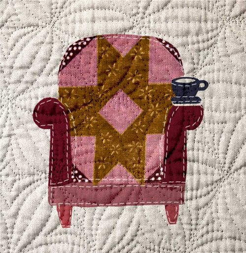 Tan quilt with a pink and yellow armchair sewn into the fibers with a mug on the arm.